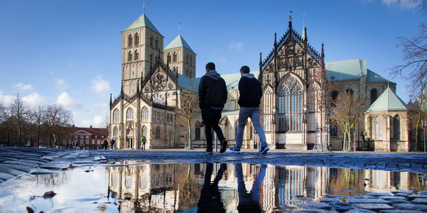 Der Dom in Münster spiegelt sich in großflächigen Pfützen, zwei Menschen schreiten über den Platz