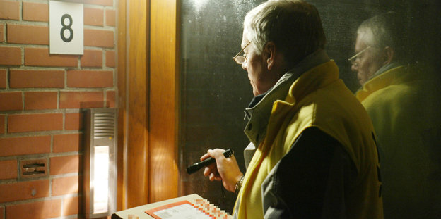 Ein Postbote mit einem Päckchen in der Hand leuchtet mit einer Taschenlampe auf eine Hausklingel