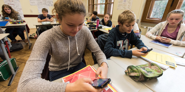 Junge Gymnasiasten sitzen in einem Klassenzimmer und tippen auf Smartphones.