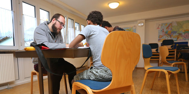 Ein junger Mann mit Bart und Brille arbeitet mit zwei Jugendlichen an einem Tisch. Alle lösen eine Aufgabe.