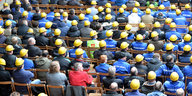 Etliche Mitarbeiter der Meyer-Werft sitzen in Arbeitskleidung auf Stühlen und blicken auf ein Podium.
