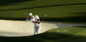 Ein Golfspieler in weißer Kleidung steht in einem Sandloch mitten in einer grünen Wiese und schlägt einen Ball ab