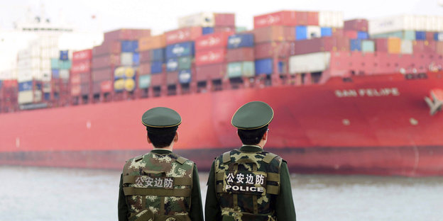 Zwei chinesische Grenzpolizisten stehen vor am Hafen vor einem großen Containerschiff