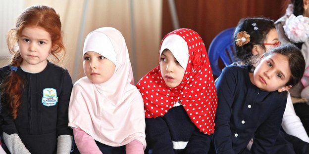 Vier kleine Mädchen im Vorschulalter sitzen nebeneinander, die zwei in der Mitte tragen Kopftücher