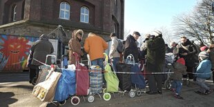 Menschen warten mit Einkaufsbeuteln vor einem Gebäude auf die Öffnung der Tafel