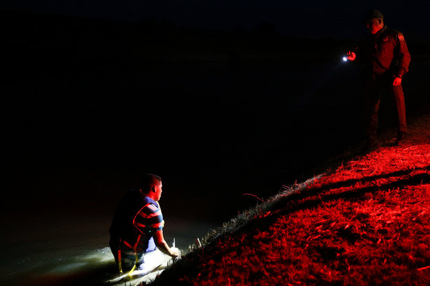 Mann kniet im Zwielicht im Wasser an der US-mexikanischen Grenze, ein Grenzpolizist leuchtet mit einer Taschenampe auf ihn.