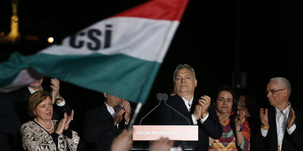 Orban klatscht in die Hände, neben ihm eine Flagge
