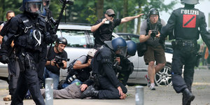 Zwei Polizisten mit Helmen hocken auf einem Demonstranten und drücken ihn mit den Knien zu Boden.