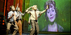 Drei Schauspieler mit Masken ziehen vor eine Leinwand mit der Großaufnahme einer Frau.