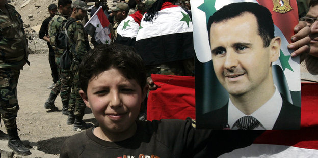 Ein Junge hält ein Assad-Portrait hoch, im Hintergrund Soldaten