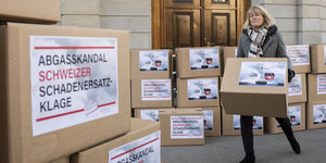 Eine Frau trägt eine Box mit der Aufschrift "Abgasskandal Schweizer Schadenersatzklage", es stehen sehr viele Boxen herum