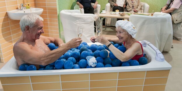 Eine Seniorin und ein Senior sitzen gemeinsam in einer Badewanne voller Plastikbälle.