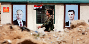 Ein Soldat läuft an einem Haus entlang. Daran kleben Poster mit den Portraits von Putin und Assad