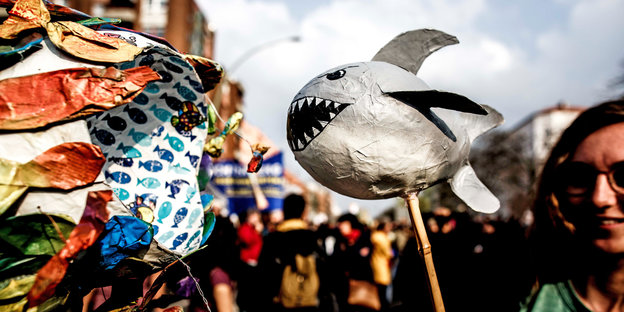 Eine Demonstrantin hält eine Pappmasché-Figur aus kleinen Fischen, die einen großen fangen, in der Hand