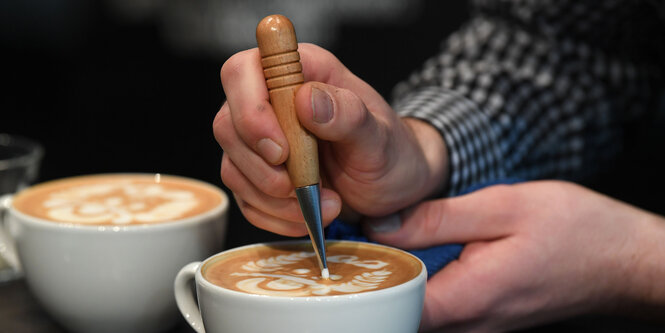 Jemand hat zwei Tassen Milchkaffee vor sich stehen und malt im Schaum der einen Tasse herum