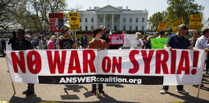 Im Hintergrund das Weiße Haus, im Vordergrund Menschen, die ein Plakat hochhalten, auf dem steht „No War on Syria“