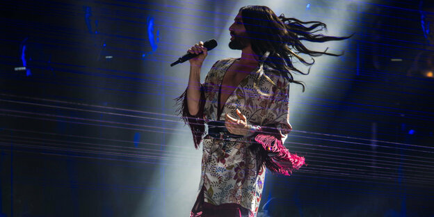 Ein Mensch mit langen Haaren sing auf einer Bühen in ein Mikrofon