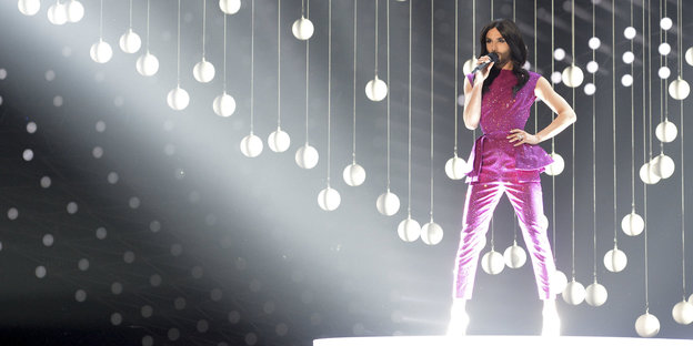 Ein Mann, der als Frau geschminkt ist und einen pinken Anzug anhat, singt auf einer beleuchteten Bühne