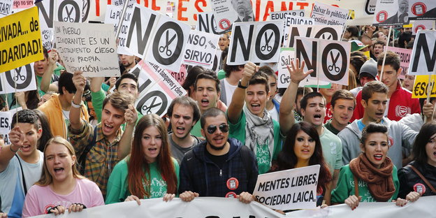Junge spanische Leute halten auf einer Demonstration Plakate und Schilder hoch, auf denen vielfach "No" und eine Schere steht.
