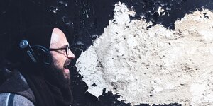 Ein Mann mit Kopfhörern steht mit geöffetem Mund vor einer Wand, deren Putz eine Sprechblase bildet