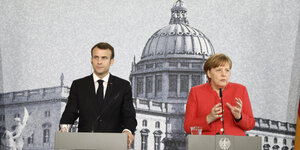 Macron im schwarzen Jackett, Merkel im roten, stehen vor einer Fototapete