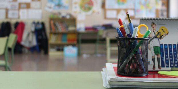Ein Behälter mit Stiften und Schere steht auf einem Tisch in einem Klassenzimmer.