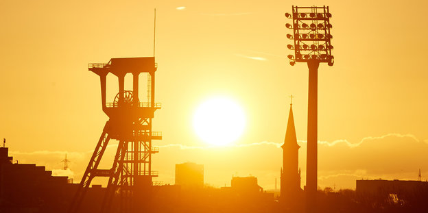 Die Sonne geht über dem Förderturm einer Zeche und einem Flutlichtmast eines Stadion auf