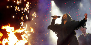 Conchita Wurst hält ein Mikrofon in der Hand und singt, neben ihr lodert ein Feuer