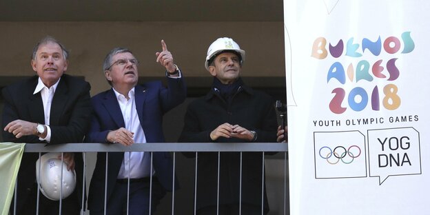 Drei Männer stehen auf einer Baustelle, neben ihnen hängt ein Plakat der Olympischen Jugendspiele