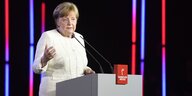 Eine Frau in weißer Bluse spricht in Mikrofone auf einem Podium, im Hintergrund bunte Lichtstreifen