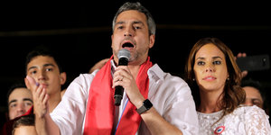 Ein Mann in einem weißen Hemd und einem roten Schal spricht in ein Mikrofon, hinter ihm eine Frau und weitere Menschen