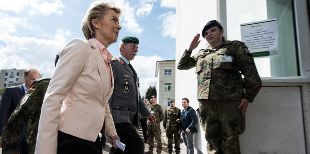 Eine Frau in Anzug geht mit uniformierten Soldaten an einer uniformierten Soldatin in ein Gebäude