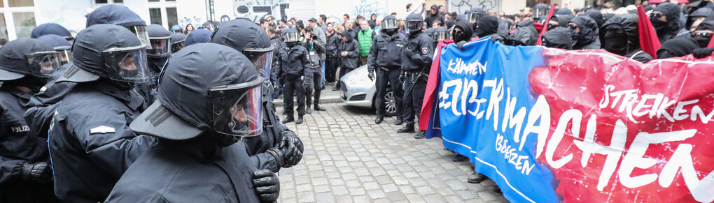 Demonstration, links die Polizei, rechts schwarz angezogene Demonstrierende, die ein Banner hochhalten