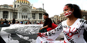 Eine in weiß gekleidete Frau trägt Schriftzüge und rote Handabdrücke auf ihrer Kleidung. Im Hintergrund eine Demonstration.