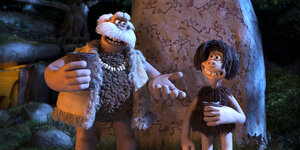 Zwei Knetfigur-Menschen, einer groß und mit weißem Schnurrbart, einer klein und mit schwarzen Haaren, stehen vor einem Stein mit Bemalungen. Sie tragen Felle