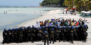 Uniformierte und Touristen stehen sich an einem Sandstrand mit Palmen gegenüber