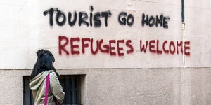 Eine Frau läuft an einer Wand vorbei, an die jemand "Tourists go home" und "Refugees Welcome" gesprüht hat