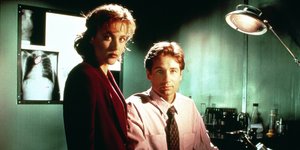 Dana Scully und Fox Mulder aus der Serie "Akte-X" blicken in die Kamera. Im Hintergrund hängen Röntgenbilder