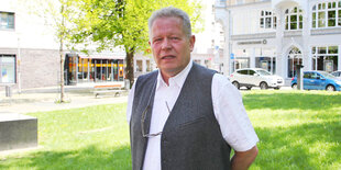 Ein älterer Mann in weißem Tshirt und grauer Weste steht auf einer grünen Wiese. Die Sonne scheint