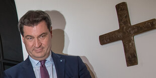 Ein Mann, Markus Söder, schaut direkt in die Kamera, neben ihm hängt ein Kreuz an der Wand