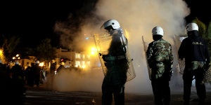 Polizisten mit weißen Helmen stehen nachts vor aufsteigendem Rauch, im Hintergrund sind Palmen und noch mehr Polizisten zu sehen