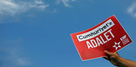 eine Hand hält ein rotes Pappschild, auf dem auf türkisch „Gerechtigkeit für cumhuriyet“ steht in den Himmel
