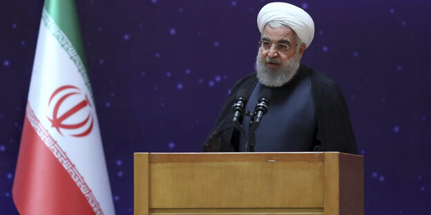 Ein Mann mit einem Turban auf dem Kopf spricht an einem Podium, links von ihm die iranische Flagge mit den Farben grün-rot-weiß