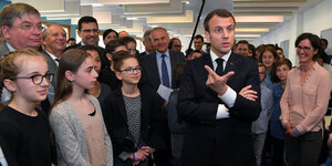 Emmanuel Macron zeigt mit den Fingern zwei Optionen an, um ihn herum stehen Jugendliche und Erwachsene