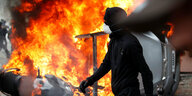 Vermummter Mann in scharz vor einem lichterloh brennenden Auto
