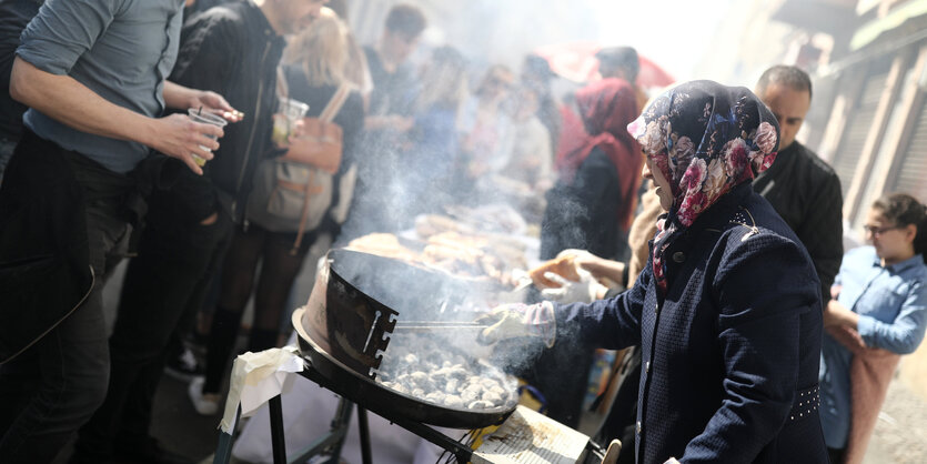 Frau brät auf einem Grill, vor ihr stehen Menschen