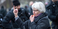 Eine Frau mit weißen Haaren bläst in eine Trillerpfeife und zeigt den Mittelfinger, im Hintergrund sieht man Polizei