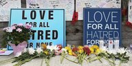 Plakate mit der Aufschrift "Love for All, Hatred for no one" lehnen an einer Mauer, davor wurden Blumen niedergelegt