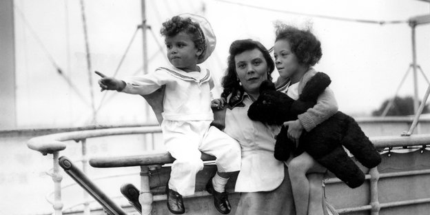 Ein Foto in schwarz weiß zeigt eine junge Frau mit einem Jungen und einem Mädchen auf dem Arm an Bord eines Schiffes. Der Junge trägt einen Matrosenanzug, das Mädchen ein Kleid und ein Plüschtier