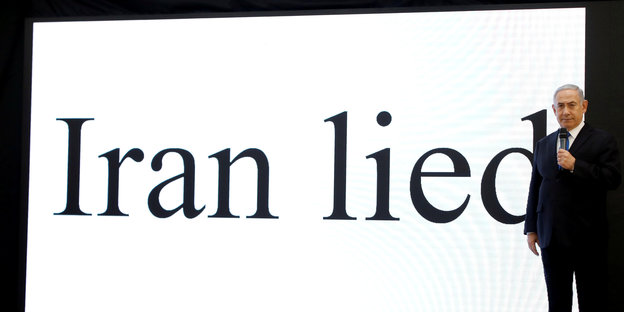 Ein Mann mit grauen Haaren steht vor einer Leinwand, auf der wird in großen Buchstaben angestrahlt: "Iran lied"